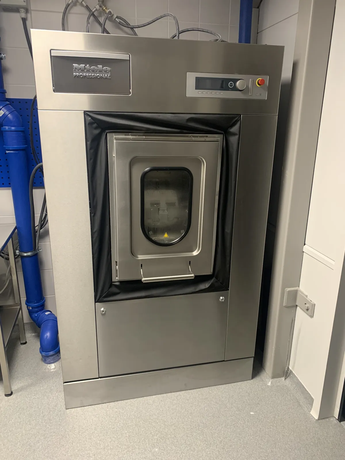 Miele Professional industritvättmaskin installerad i ett tvättrum med blå tilloppsledningar