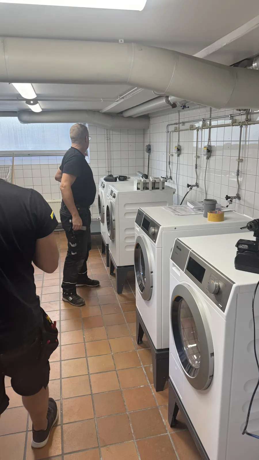 Två tekniker som arbetar med tvättmaskiner i en källartvättstuga med klinkergolv